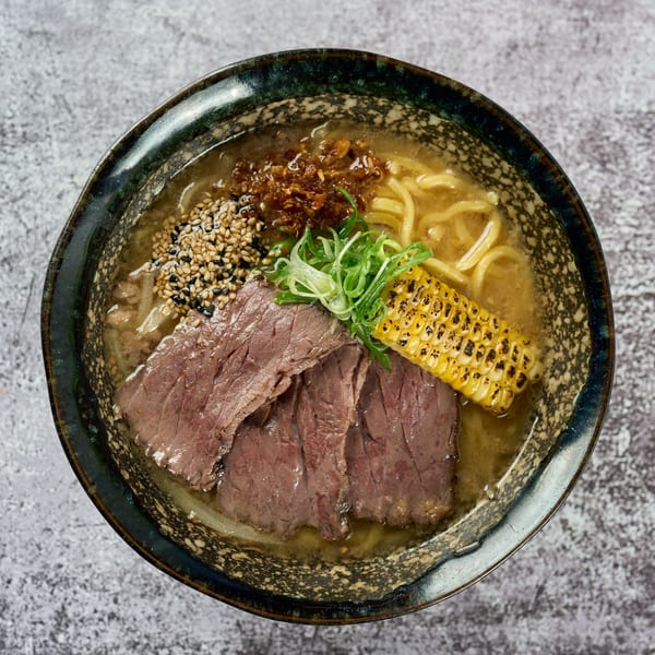 Overhead view of KEI ramen with beef, grilled corn, green onions, and sesame seeds in a dark bowl.