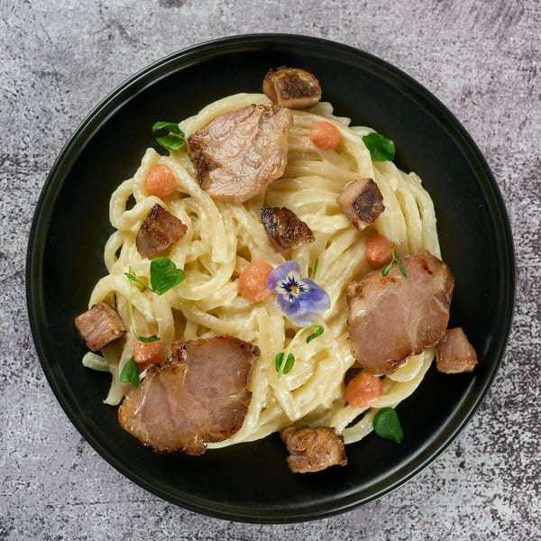 Creamy Japanese udon noodles with pork belly, orange spheres, and a purple edible flower.