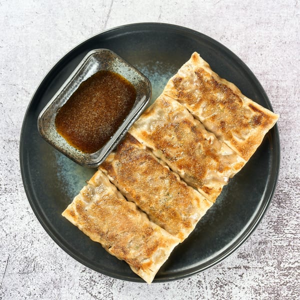 Four pan-fried gyoza dumplings with a side of dipping sauce on a dark plate.