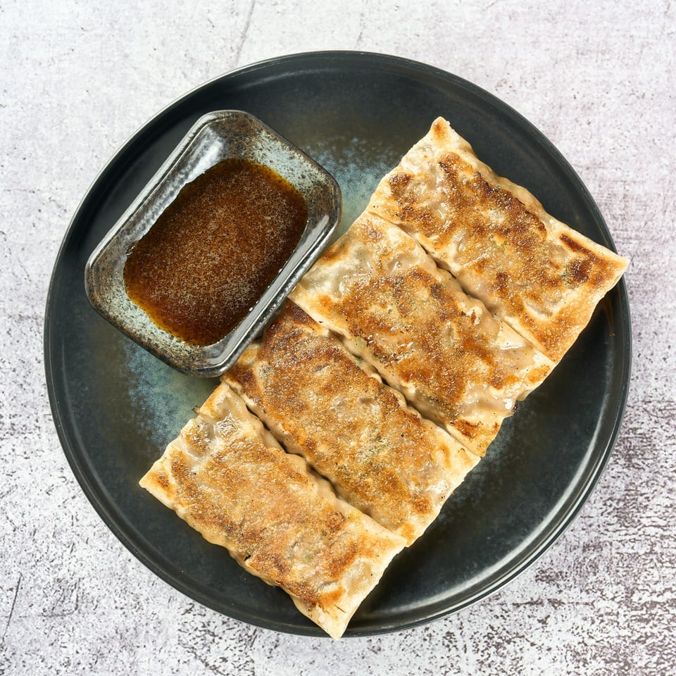 Four pan-fried gyoza dumplings with a side of dipping sauce on a dark plate.