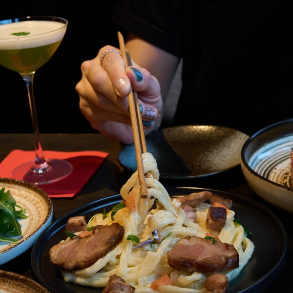 Chopsticks lifting creamy udon noodles with duck slices and a green cocktail on a dark table.